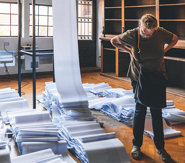 A male worker is looking at a stack of newly woven fabric in possibly a mill, the fabric is draped from the ceiling